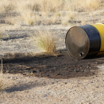 A tipped oil barrel spills its contents onto the soil in an open field.